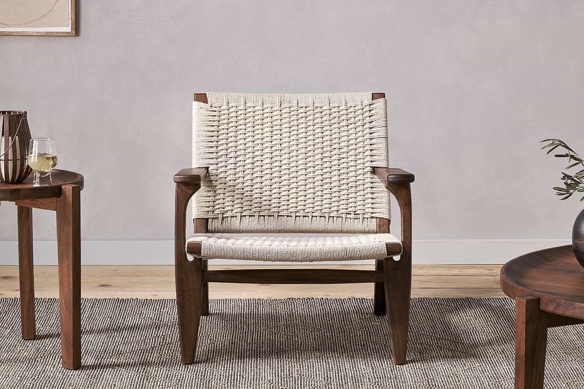 The Surat Woven Lounger - Natural by nkuku features acacia wood, wide armrests, and a hand-woven cotton rope seat and back. Placed on a textured gray rug, it sits between two round tables—one with white wine, the other with a vase of greenery.