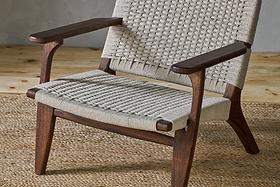 The Surat Woven Lounger - Natural by nkuku features angled acacia wood legs and a hand-woven cotton rope seat. Wide, flat armrests complement its timeless look, perfect over a natural fiber rug against a light gray wall.