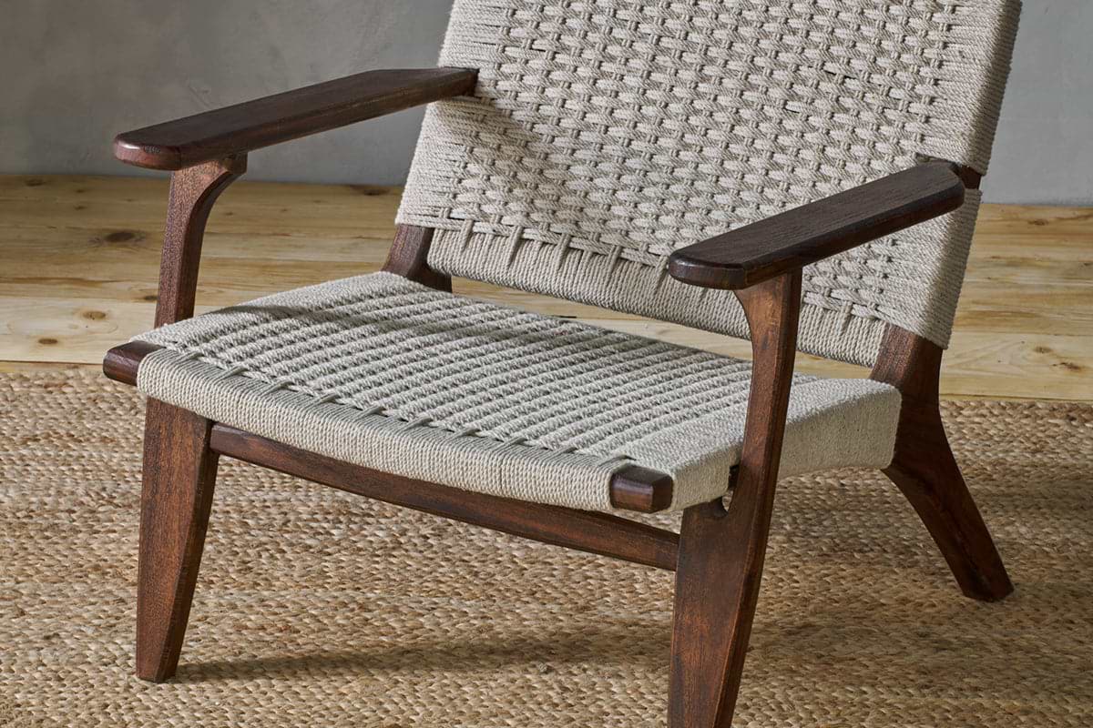 The Surat Woven Lounger - Natural by nkuku features angled acacia wood legs and a hand-woven cotton rope seat. Wide, flat armrests complement its timeless look, perfect over a natural fiber rug against a light gray wall.