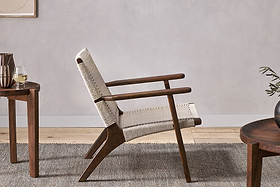 The Surat Woven Lounger - Natural by nkuku, crafted from acacia wood with a hand-woven cotton rope seat and backrest, sits on a textured gray rug beside a round wooden side table with white wine in a light, minimalist room.