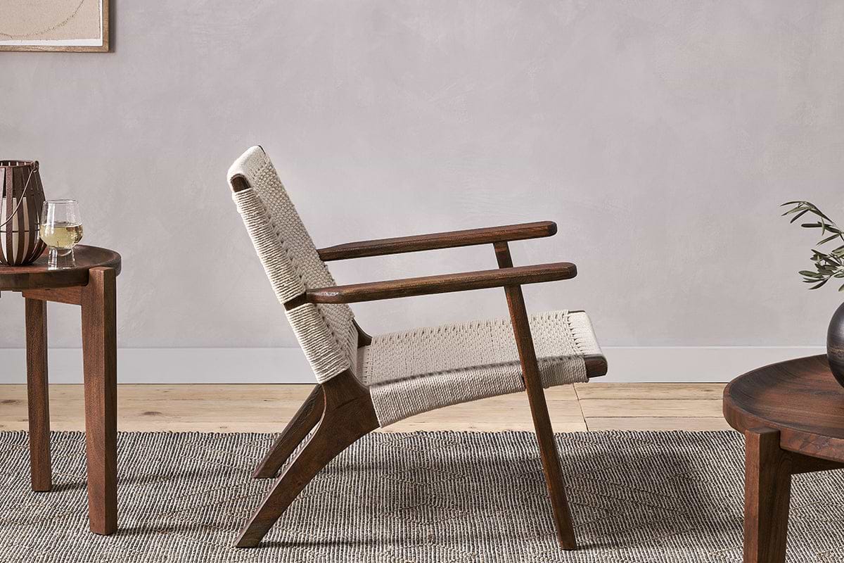 The Surat Woven Lounger - Natural by nkuku, crafted from acacia wood with a hand-woven cotton rope seat and backrest, sits on a textured gray rug beside a round wooden side table with white wine in a light, minimalist room.