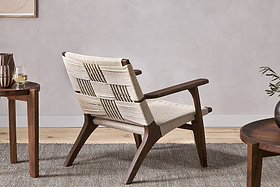 The nkuku Surat Woven Lounger - Natural, featuring hand-woven cotton rope and acacia wood, sits on a textured rug beside a small round wooden table with a glass of white wine and another table with a plant, all against a light neutral wall.