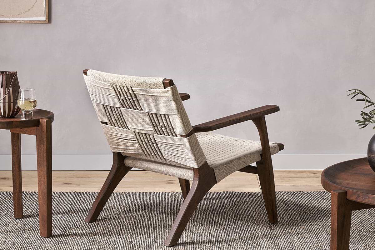 The nkuku Surat Woven Lounger - Natural, featuring hand-woven cotton rope and acacia wood, sits on a textured rug beside a small round wooden table with a glass of white wine and another table with a plant, all against a light neutral wall.