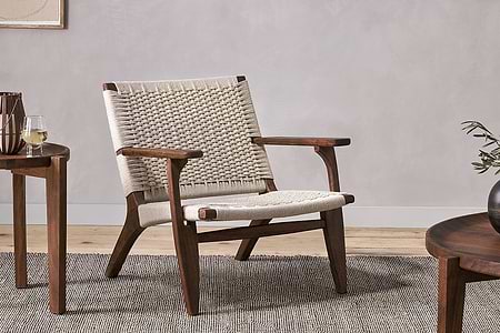 The Surat Woven Lounger - Natural by nkuku features acacia wood with a hand-woven cotton rope seat and backrest. Set on a gray rug, it’s styled with angled legs, armrests, and two wooden tables—one with a wine glass, the other with greenery.