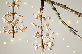 Three handmade Tanu Mango Wood Beaded Baubles by nkuku in brass and natural tones adorn a lichen-covered branch. Their elegant wood and beaded design makes them perfect, unique Christmas ornaments, softly illuminated by warm fairy lights in the background.