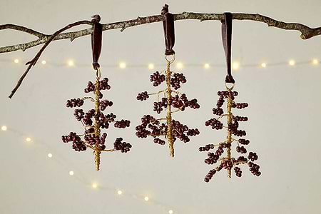 The nkuku Tanu Mango Wood Beaded Baubles - Brass & Ruby (Set of 3) feature ruby-toned and brass beads on gold wire, hanging from dark ribbons. Soft, warm lights glow in the background, adding a festive touch.