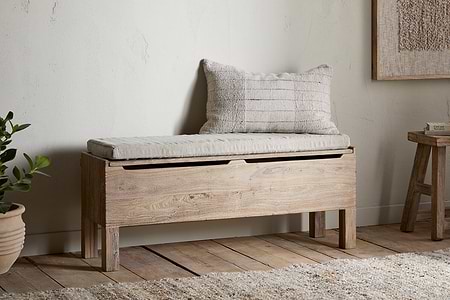 The nkuku Teni Reclaimed Wood Storage Bench with Upholstered Linen Cushion sits against a light wall. A potted plant and wooden stool rest on the floor nearby, creating a cozy entryway scene.