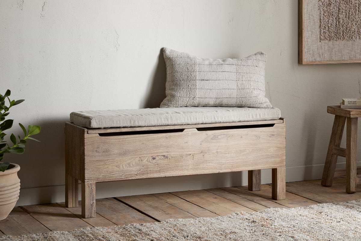 The nkuku Teni Reclaimed Wood Storage Bench with Upholstered Linen Cushion sits against a light wall. A potted plant and wooden stool rest on the floor nearby, creating a cozy entryway scene.