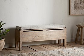 The nkuku Teni Reclaimed Wood Storage Bench with Upholstered Linen Cushion in Natural sits against a white wall on wooden floors, paired with a potted plant and wooden stool for minimalist charm.