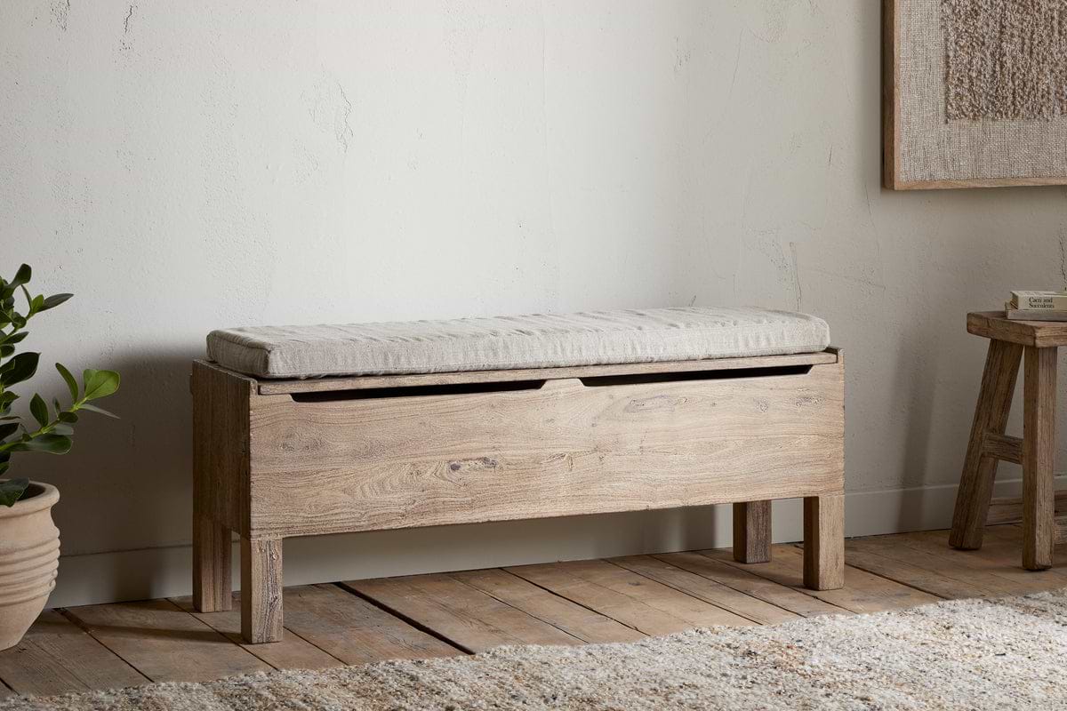 The nkuku Teni Reclaimed Wood Storage Bench with Upholstered Linen Cushion in Natural sits against a white wall on wooden floors, paired with a potted plant and wooden stool for minimalist charm.