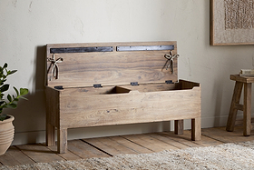 The nkuku Teni Reclaimed Wood Storage Bench with Upholstered Linen Cushion in Natural stands on wooden floors beside a potted plant and small stool, its open lid revealing interior compartments against a light-colored wall.
