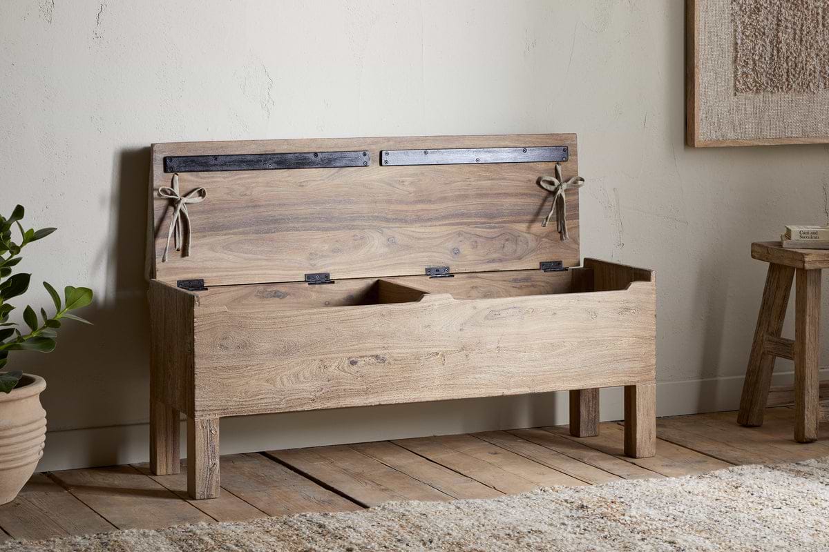 The nkuku Teni Reclaimed Wood Storage Bench with Upholstered Linen Cushion in Natural stands on wooden floors beside a potted plant and small stool, its open lid revealing interior compartments against a light-colored wall.