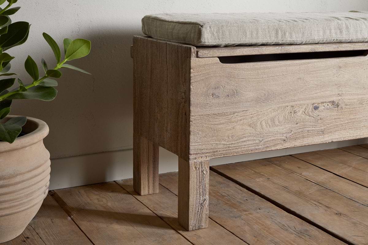 The nkuku Teni Reclaimed Wood Storage Bench with Upholstered Linen Cushion in Natural sits on a wooden floor by a potted plant. Its visible wood grain and lift-top lid offer storage, making it a stylish and practical entryway bench.