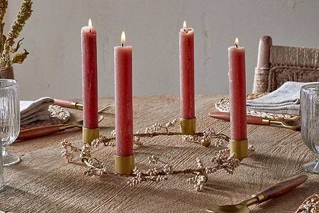 A rustic table features nkuku's Tanu Mango Wood Beaded Candle Holders in brass and natural, each with a lit red candle arranged in a circle, surrounded by a white flower garland, crystal glasses, napkins, and wooden-handled cutlery for a festive setting.
