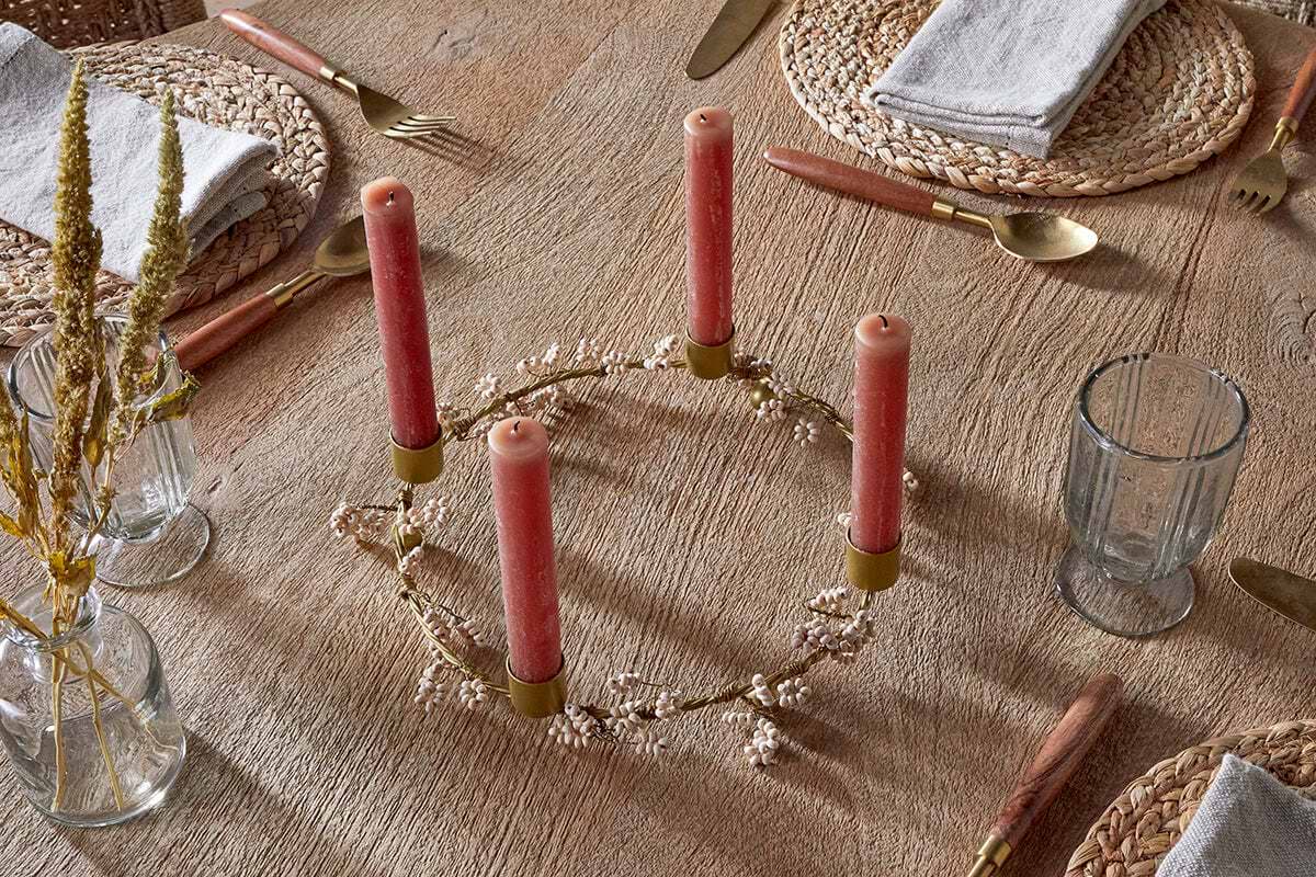 A wooden table set for a meal features woven placemats, gold cutlery, and gray napkins. At the center, nkuku’s Tanu Mango Wood Beaded Candle Holder in brass & natural holds four pink candles amid white flowers; glasses and dried blooms complete the look.