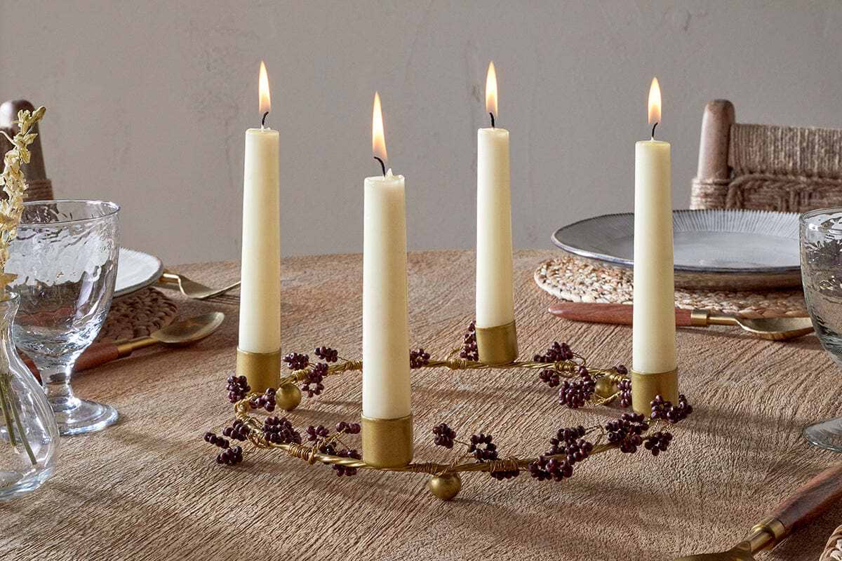 The Tanu Mango Wood Beaded Candle Holder in Brass & Ruby by nkuku, adorned with small purple berries and four tall cream candles, serves as an elegant centerpiece on a textured table set with plates, woven placemats, clear glasses, and gold utensils.