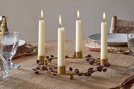 The Tanu Mango Wood Beaded Candle Holder in Brass & Ruby by nkuku, adorned with small purple berries and four tall cream candles, serves as an elegant centerpiece on a textured table set with plates, woven placemats, clear glasses, and gold utensils.