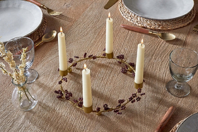 A round wooden table is set for dining, featuring the nkuku Tanu Mango Wood Beaded Candle Holder in Brass & Ruby with four cream candles and burgundy berries as a festive centerpiece. Nearby, dried flowers sit in a small vase.