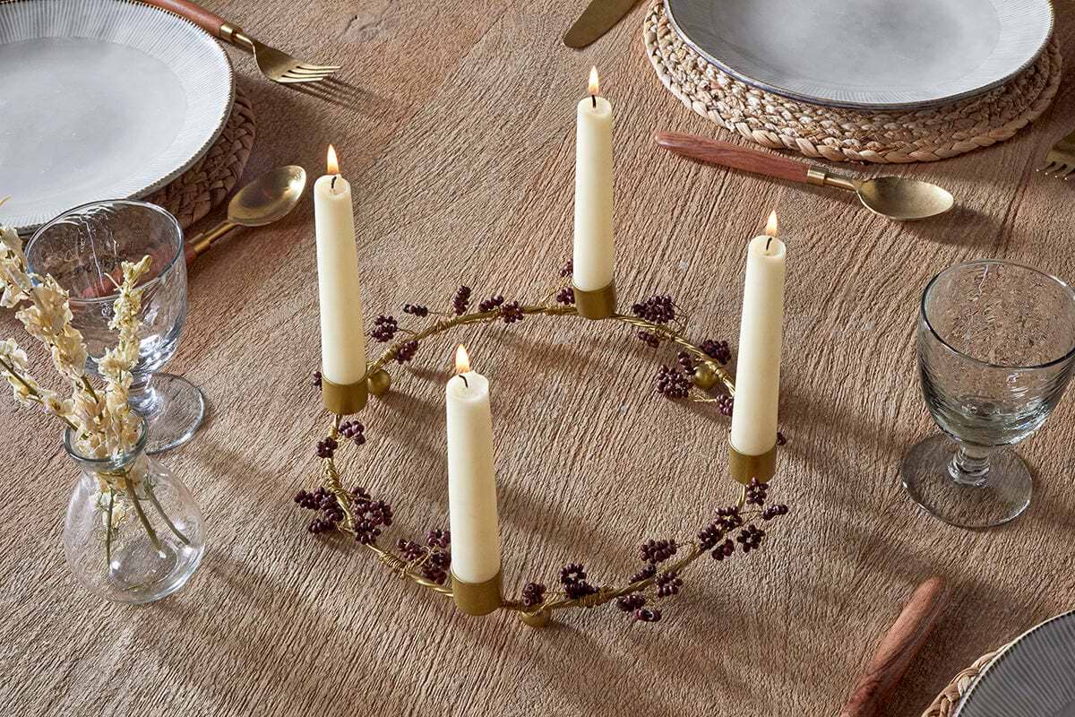 A round wooden table is set for dining, featuring the nkuku Tanu Mango Wood Beaded Candle Holder in Brass & Ruby with four cream candles and burgundy berries as a festive centerpiece. Nearby, dried flowers sit in a small vase.