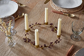 The nkuku Tanu Mango Wood Beaded Candle Holder in Brass & Ruby, styled with five cream candles and purple florals, is a stunning centerpiece for festive tablescapes. A small vase of dried flowers sits beside this elegant decorative accent.
