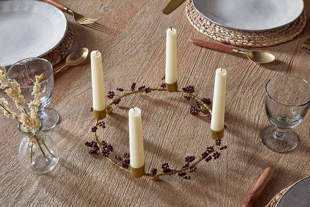The nkuku Tanu Mango Wood Beaded Candle Holder in Brass & Ruby, styled with five cream candles and purple florals, is a stunning centerpiece for festive tablescapes. A small vase of dried flowers sits beside this elegant decorative accent.