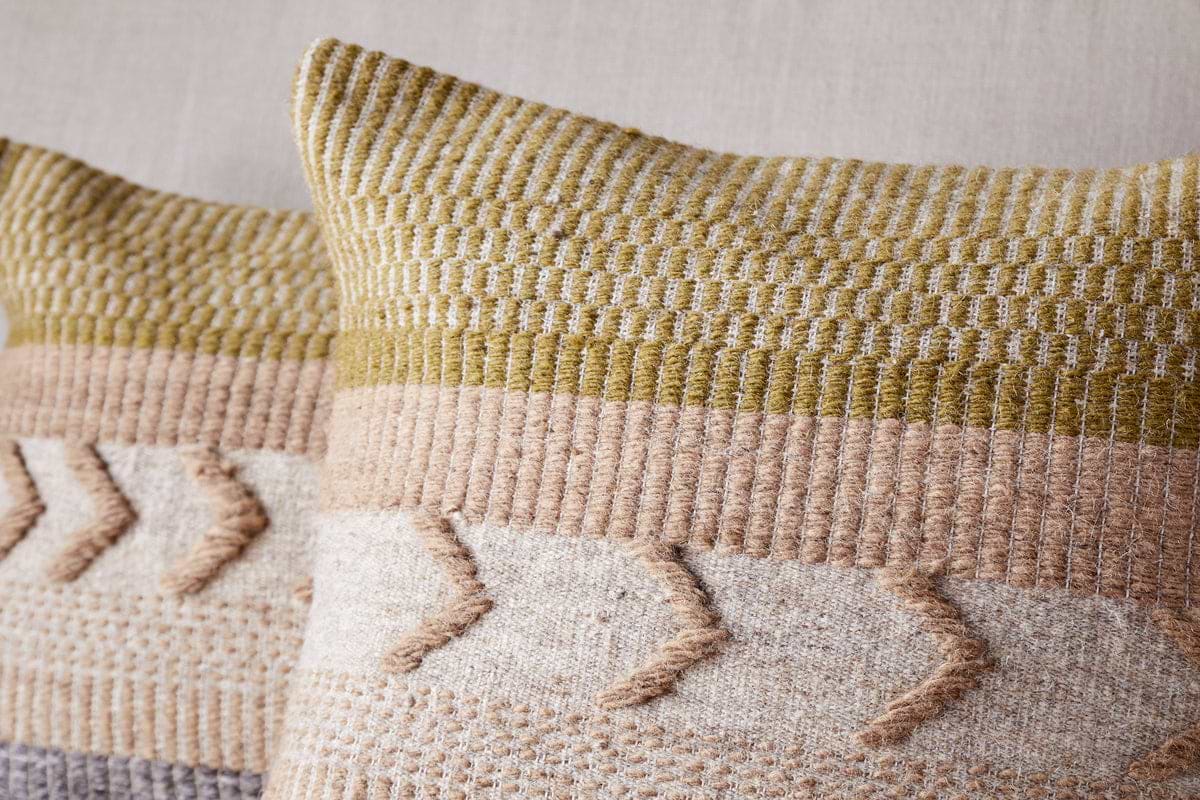 The nkuku Tuleeka Wool Cushion Cover in ochre features a textured woven design in beige, tan, and olive green with chevron-style raised details across the lower half. One decorative cover is shown prominently in the front.