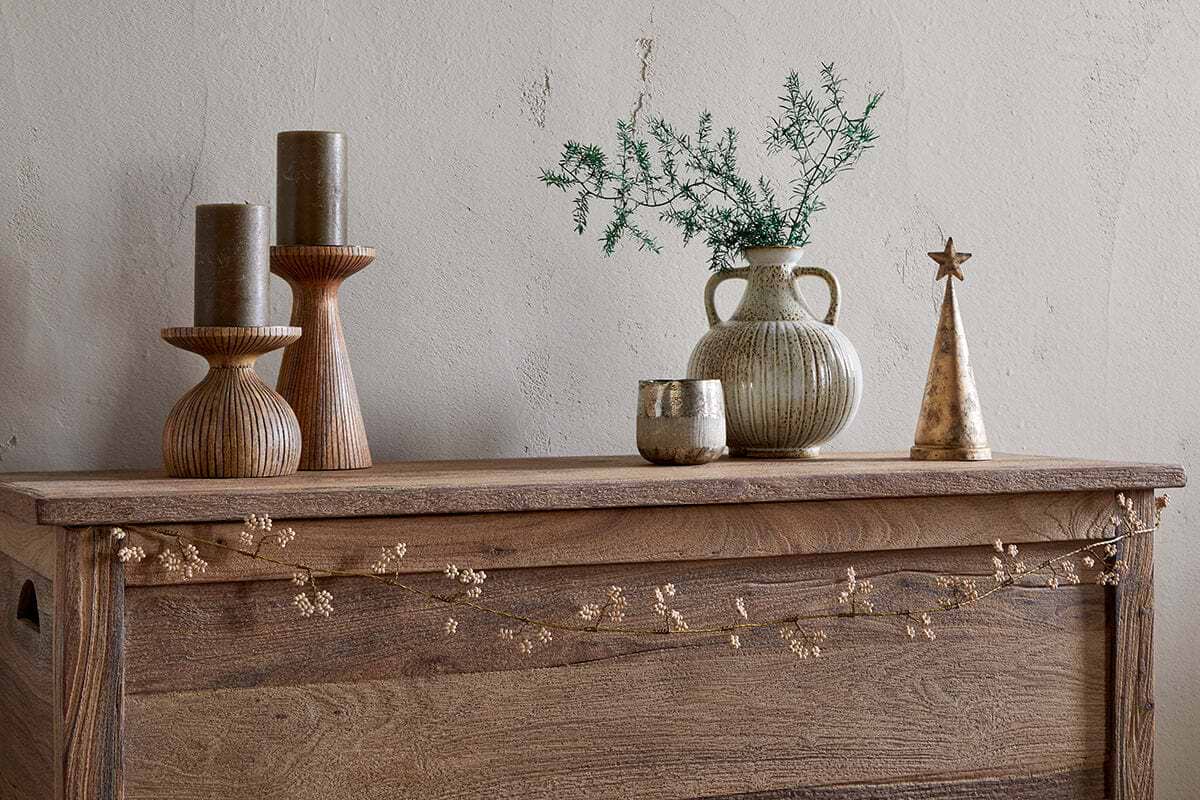 A wooden chest displays two tall ribbed candleholders with olive candles, a small ribbed vase with greenery, an antique brass votive, a gold cone-shaped tree topped with a star, and nkuku's Tanu Mango Wood Beaded Garland in Brass & Natural draped across the front.