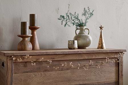 A wooden chest displays two tall ribbed candleholders with olive candles, a small ribbed vase with greenery, an antique brass votive, a gold cone-shaped tree topped with a star, and nkuku's Tanu Mango Wood Beaded Garland in Brass & Natural draped across the front.