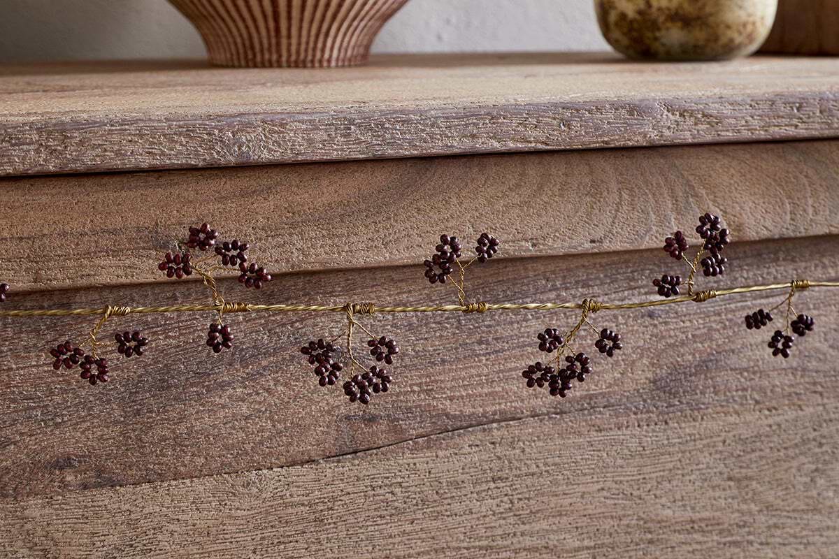 A close-up of the nkuku Tanu Mango Wood Beaded Garland in Brass & Ruby, featuring gold garland with berry-like beads on a rustic wooden surface, with blurred pottery vases in the background.