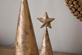 Two metallic, cone-shaped Talna Tree Bell ornaments by nkuku, with an antique brass finish, sit on a surface; the smaller one is topped with a gold star. A round textured gold decoration hangs behind, adding festive charm to your decor.