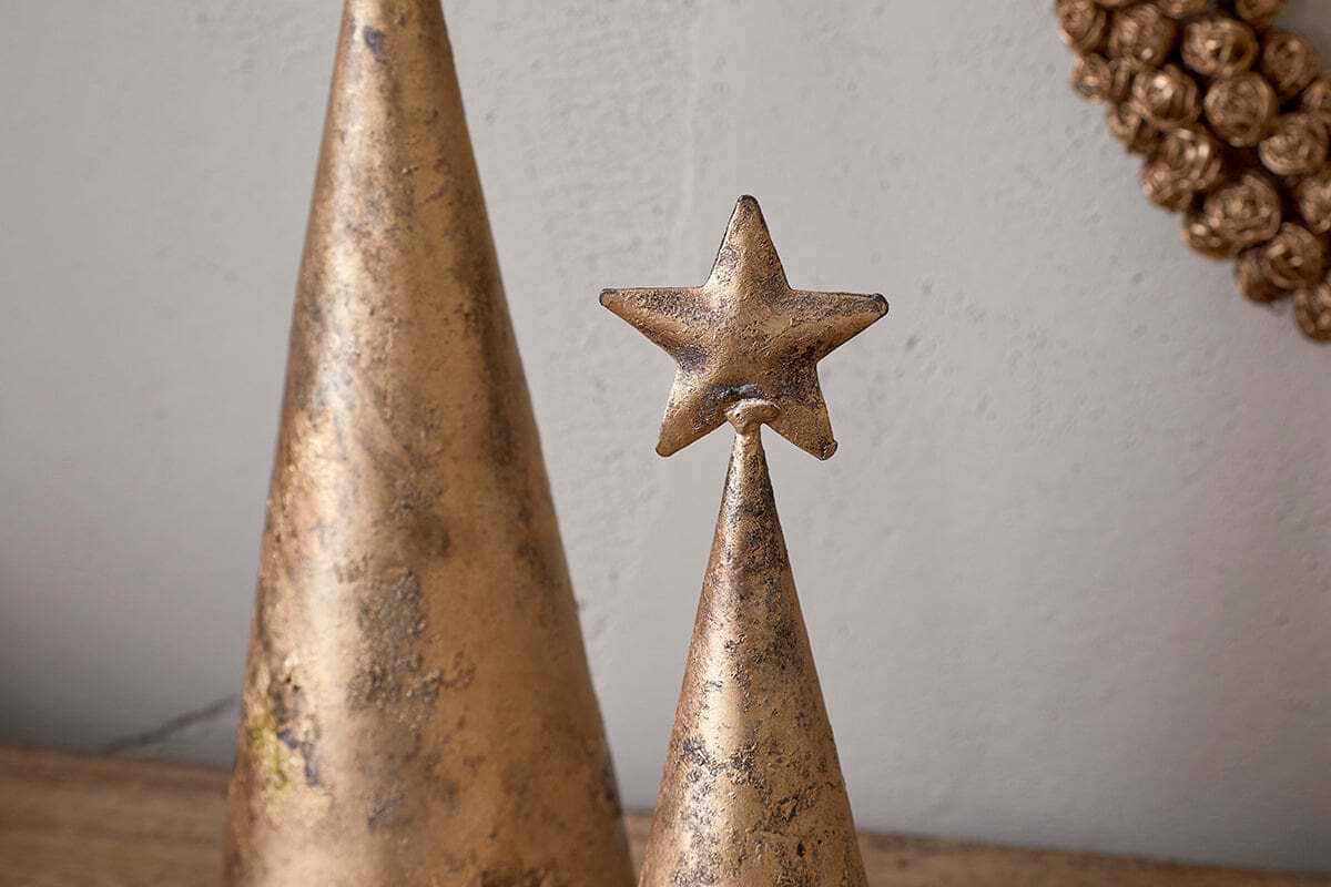 Two metallic, cone-shaped Talna Tree Bell ornaments by nkuku, with an antique brass finish, sit on a surface; the smaller one is topped with a gold star. A round textured gold decoration hangs behind, adding festive charm to your decor.