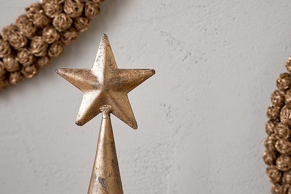 A close-up of the nkuku Talna Tree Bell in antique brass showcases its festive charm on a cone-shaped base. The background has a plain, light wall with decorative, braided wreaths partially visible on each side.