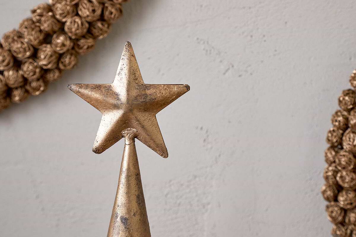 A close-up of the nkuku Talna Tree Bell in antique brass showcases its festive charm on a cone-shaped base. The background has a plain, light wall with decorative, braided wreaths partially visible on each side.