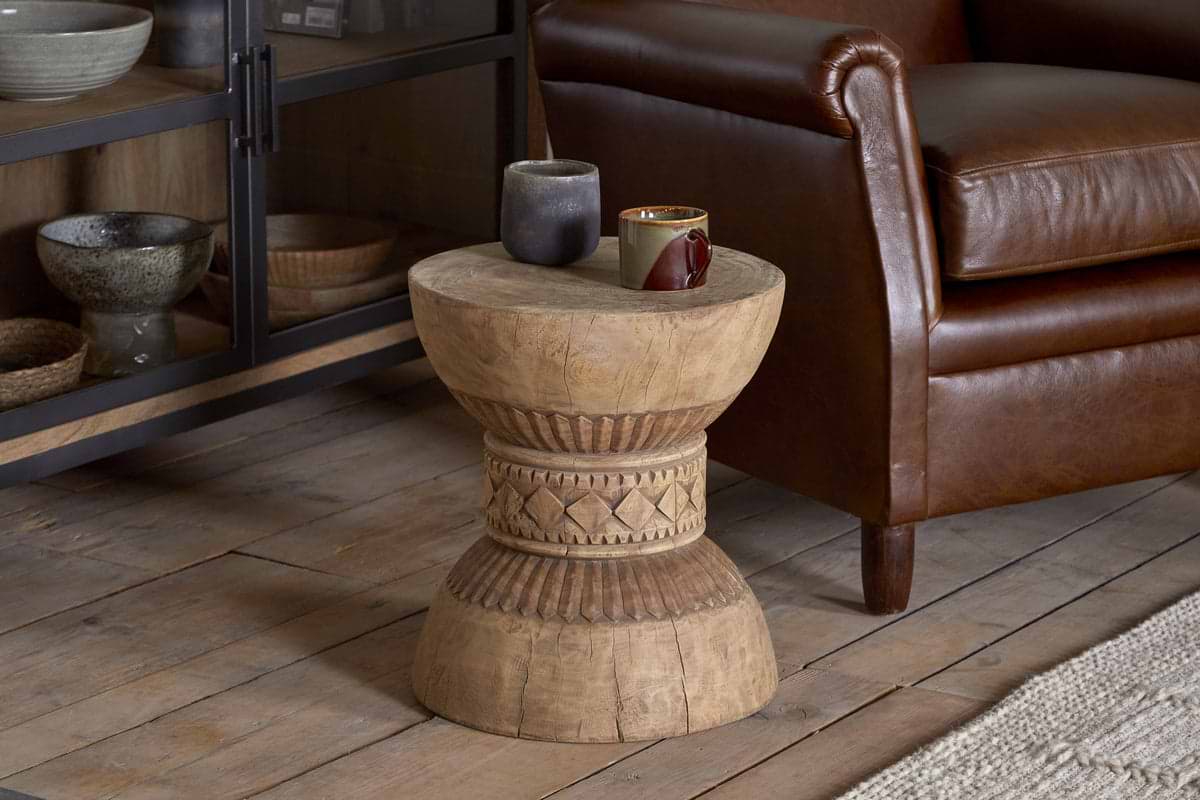 The Taru Carved Side Table - Natural by nkuku, featuring intricate hand-carved patterns, stands on a wooden floor beside a brown leather armchair. Two mugs and a small container are placed on this Indian-inspired piece, with shelves of bowls behind.