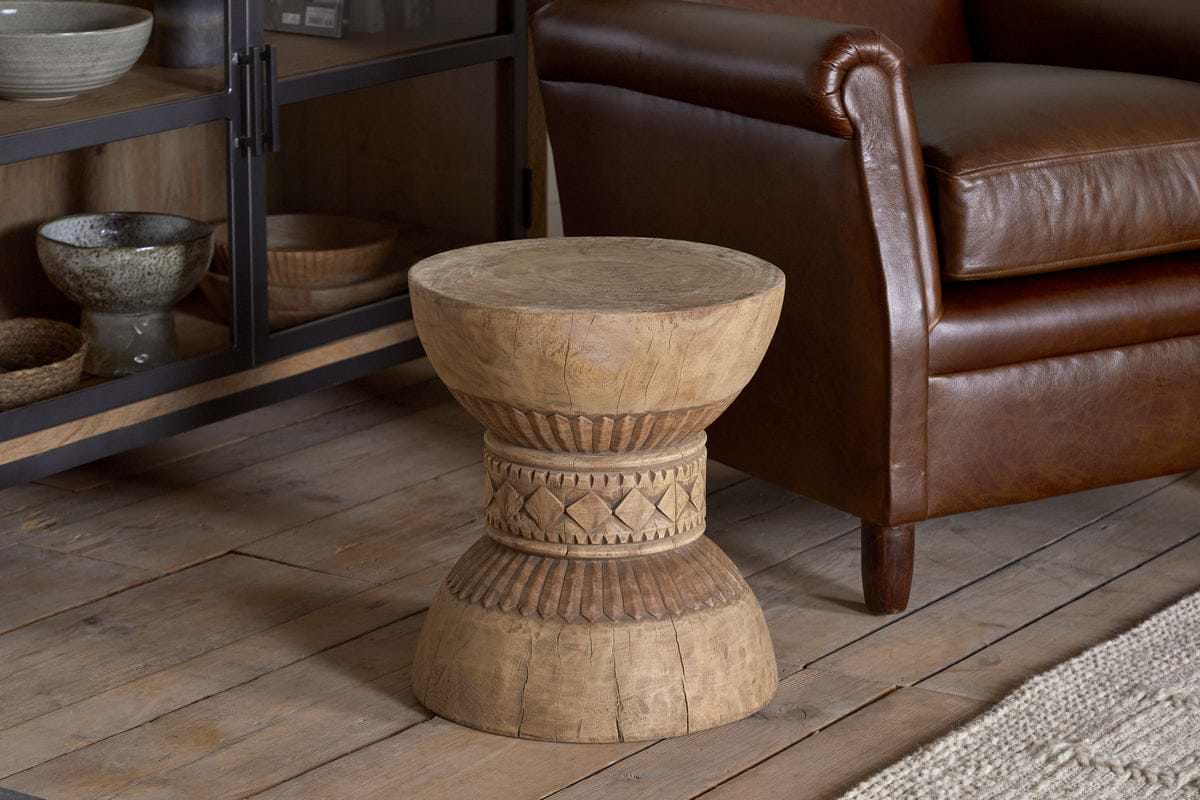 The Taru Carved Side Table - Natural by nkuku, featuring intricate geometric patterns, rests on a wooden floor beside a brown leather armchair and a glass-fronted cabinet filled with bowls.