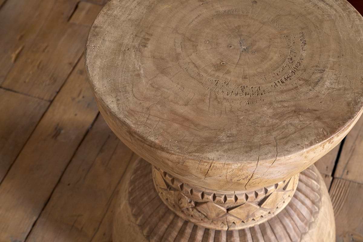 A close-up of the Taru Carved Side Table - Natural by nkuku, a round rustic solid wood piece featuring decorative carved patterns on its base, shown on a wooden floor with visible grain.