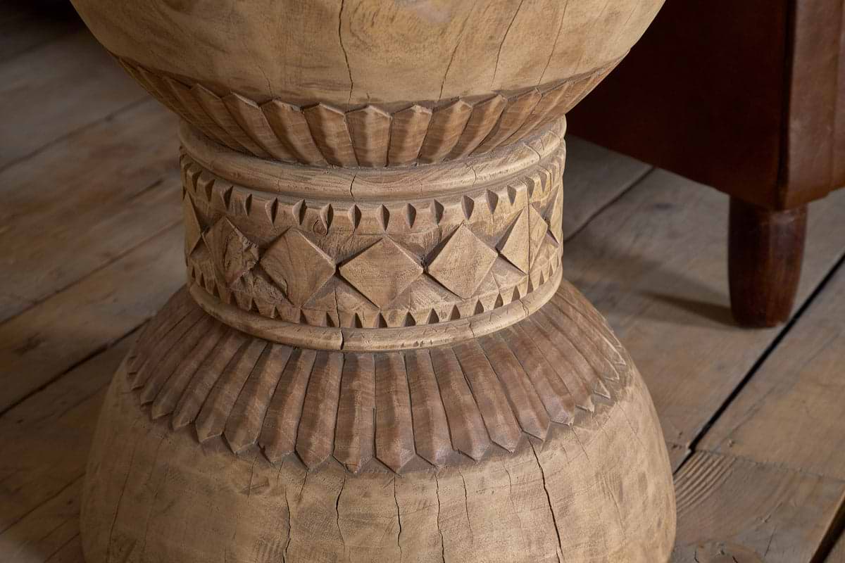 Close-up of the Taru Carved Side Table - Natural by nkuku, featuring a hand-carved leg with intricate diamond and vertical line patterns inspired by Indian design, set on a wooden floor.