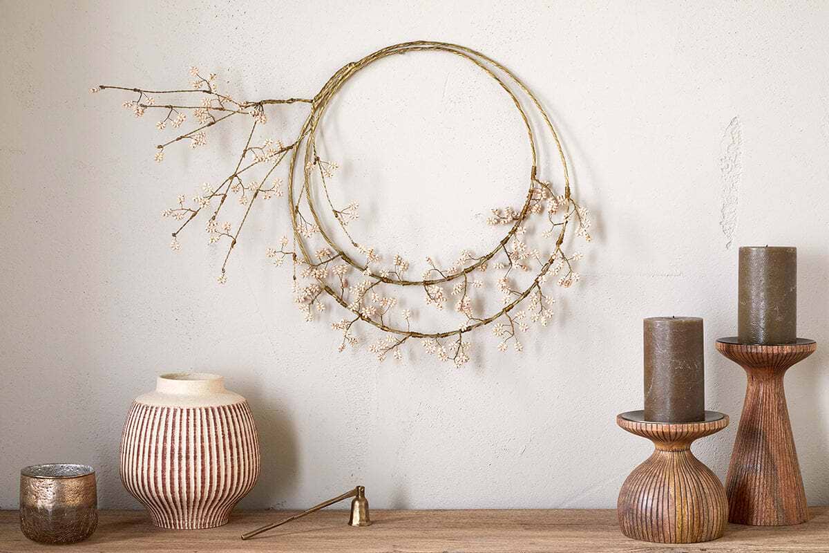 A minimalist, neutral tabletop features ribbed ceramic vases, dark pillar candles on wood stands, a brass candle snuffer, and nkuku’s Tanu Layered Mango Wood Beaded Wreath in Brass & Natural, adorned with white faux flowers and branches.