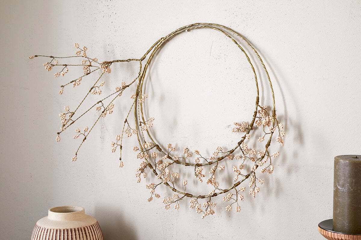 The Tanu Layered Mango Wood Beaded Wreath by nkuku, featuring brass and natural wood beads, hangs on a light textured wall. A ceramic vase and a dark cylindrical object are placed nearby for a stylish minimal look.