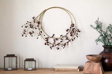 The nkuku Tanu Layered Mango Wood Beaded Wreath in Brass & Ruby hangs on a cream wall. Below, square lanterns, a brown vase with a plant, a stacked wooden bowl, and a book form an elegant festive centrepiece.