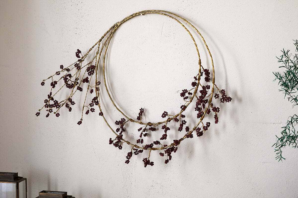 The Tanu Layered Mango Wood Beaded Wreath by nkuku features two slim brass rings with ruby red beads, hanging on a light textured wall. Green foliage appears on the right side of the image.