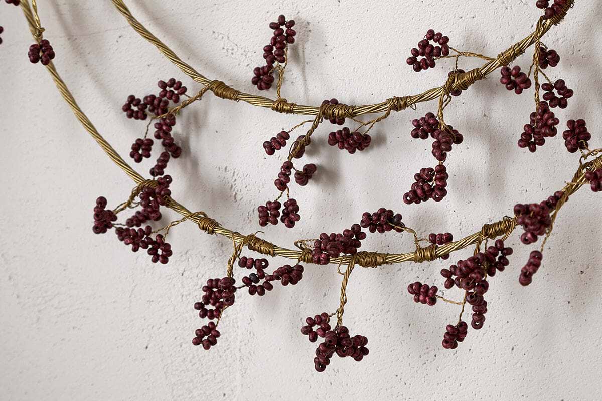Close-up of two nkuku Tanu Layered Mango Wood Beaded Wreaths in Brass & Ruby, featuring twisted, branch-like designs and clusters of rich ruby beads for a rustic, antique-inspired look against a white textured wall.