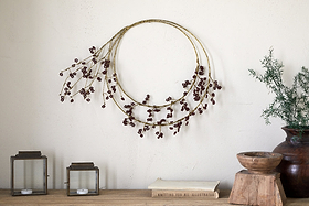 The nkuku Tanu Layered Mango Wood Beaded Wreath in brass and ruby hangs above a wooden surface. Below, antique brass accents—glass lanterns, books, a wooden bowl, and a dark vase with greenery—create a festive centerpiece.