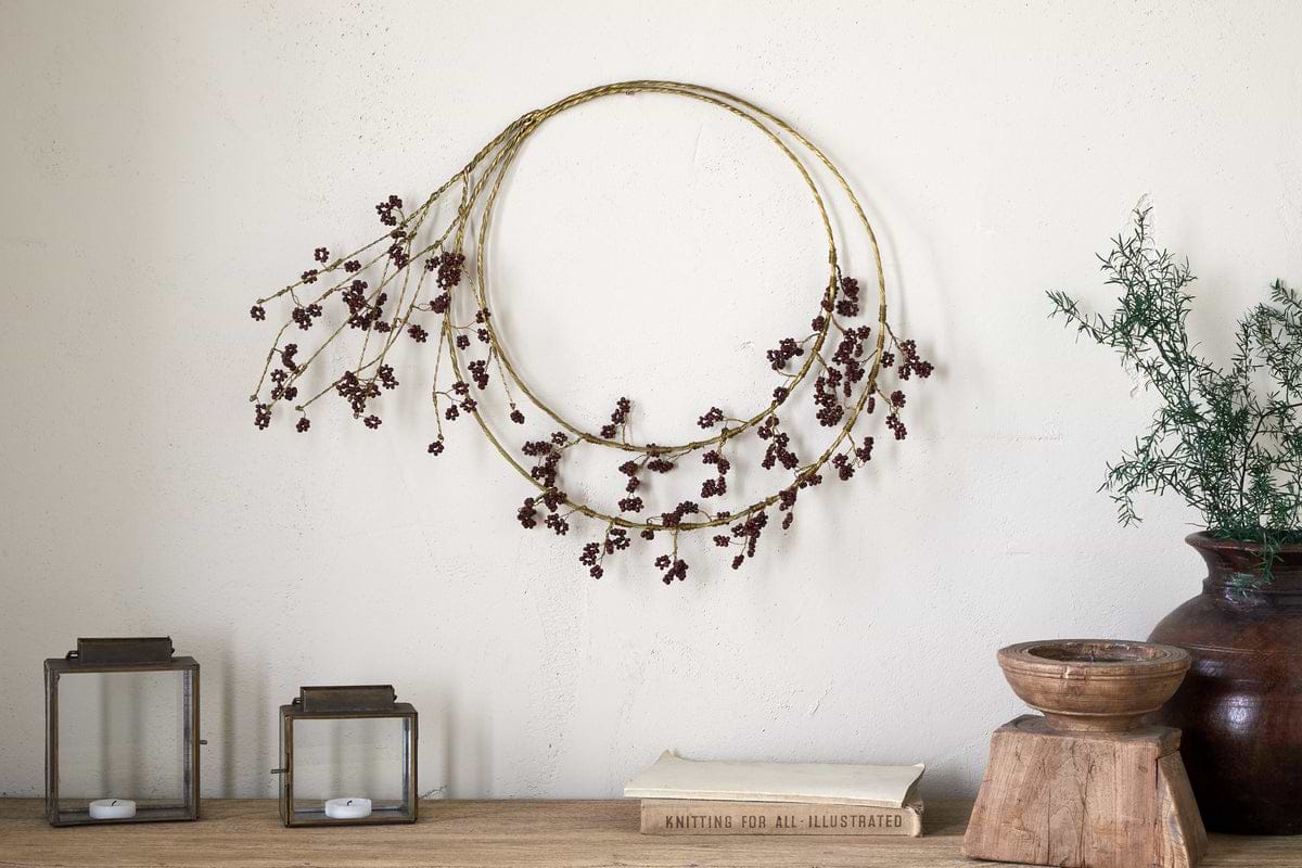 The nkuku Tanu Layered Mango Wood Beaded Wreath in brass and ruby hangs above a wooden surface. Below, antique brass accents—glass lanterns, books, a wooden bowl, and a dark vase with greenery—create a festive centerpiece.
