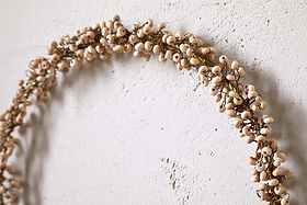 The nkuku Tanu Mango Wood Beaded Wreath in brass and natural features minimalist wooden beads for a rustic Christmas touch, shown hanging on a textured off-white wall.