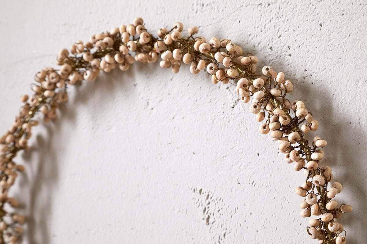 The nkuku Tanu Mango Wood Beaded Wreath in brass and natural features minimalist wooden beads for a rustic Christmas touch, shown hanging on a textured off-white wall.