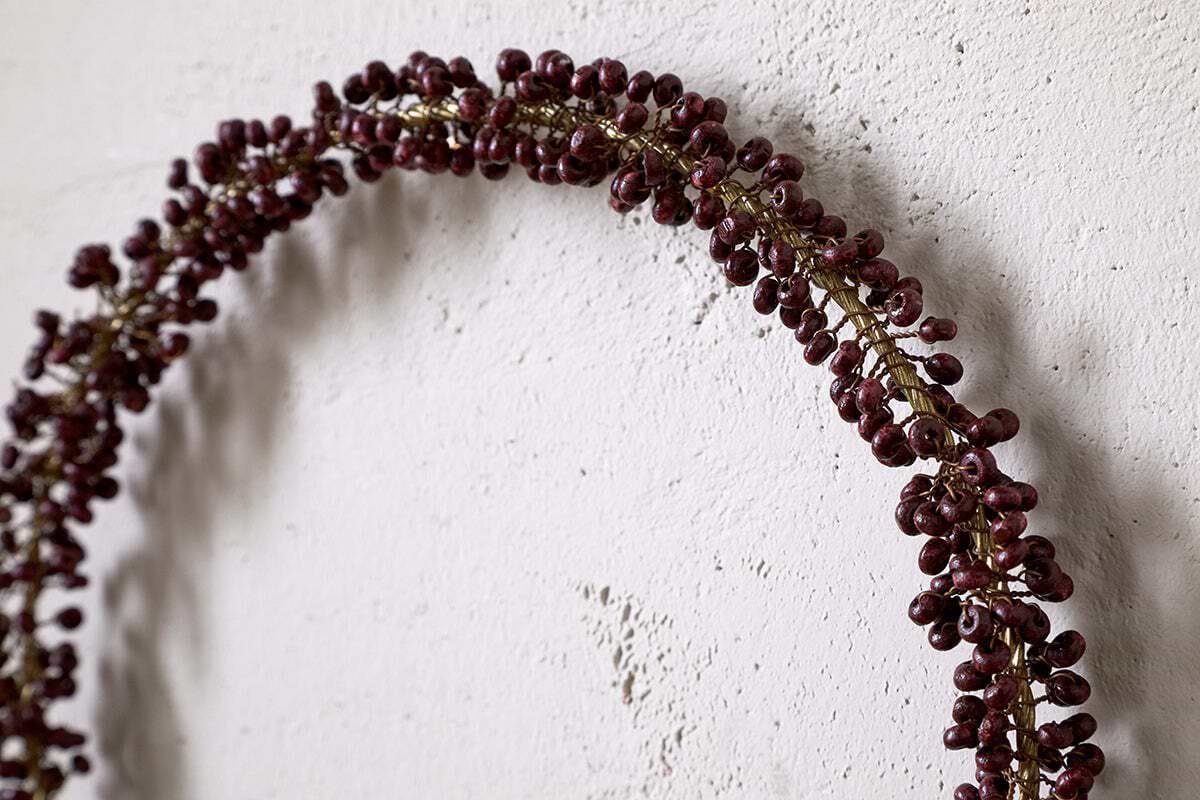 A partial view of nkuku's Tanu Mango Wood Beaded Wreath in Brass & Ruby, featuring wood beads and deep red accents, hangs on a textured white wall—ideal for Christmas decor.