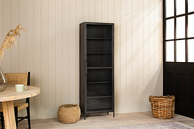 The Takua Iron Cabinet - Narrow by nkuku stands against a beige paneled wall. Nearby, a round wooden table with a mug, woven chair, and two wicker baskets rest on a light wood floor by a large window and black door. Dried grass decorates the table.