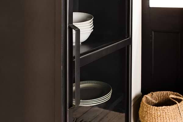 Close-up of the Takua Iron Cabinet - Narrow by nkuku, featuring a black glass front and metal handle. White bowls are on the top shelf and plates on the lower, with a woven basket nearby beside a black door with a white window.