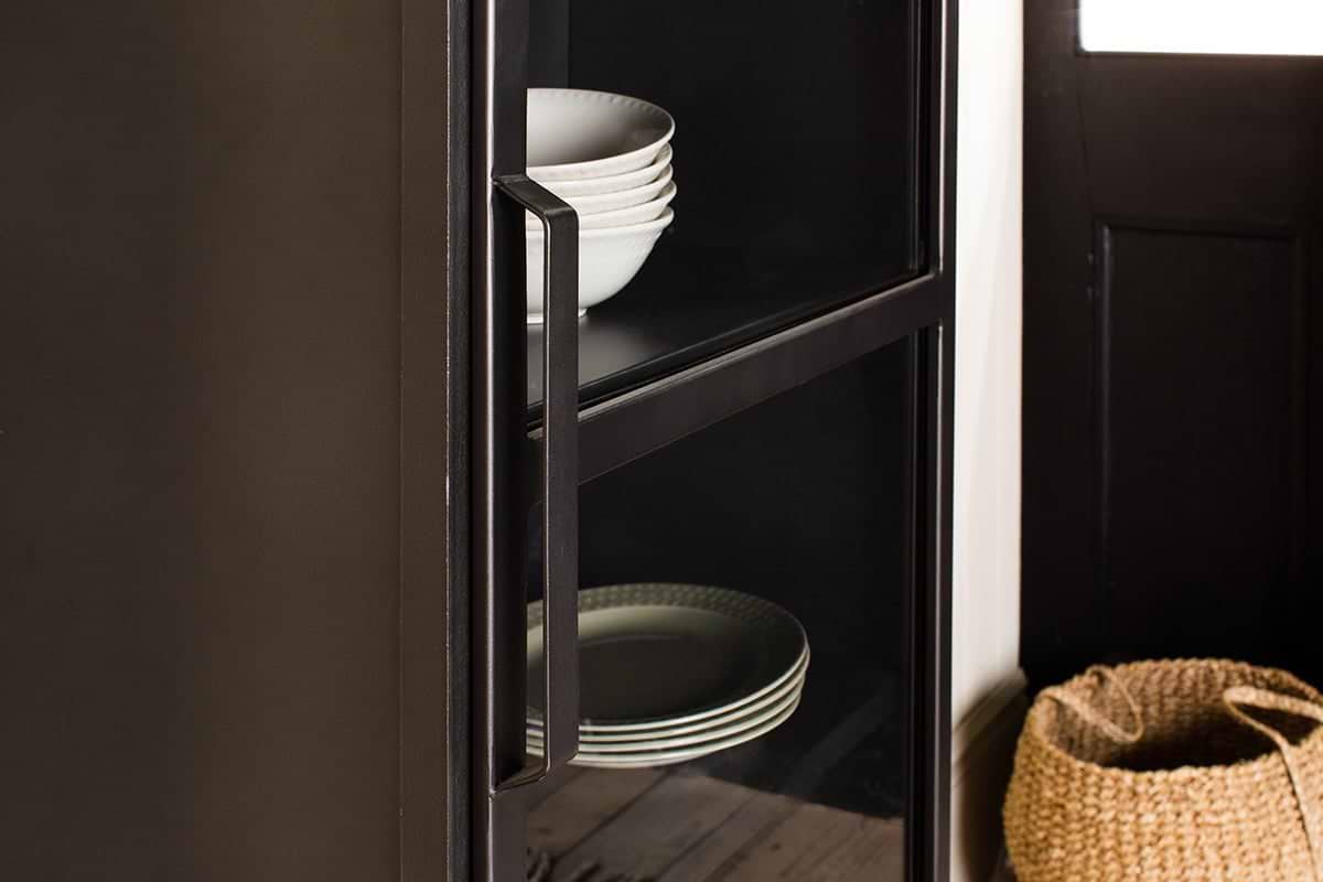 Close-up of the Takua Iron Cabinet - Narrow by nkuku, featuring a black glass front and metal handle. White bowls are on the top shelf and plates on the lower, with a woven basket nearby beside a black door with a white window.